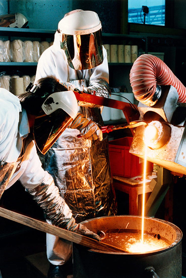 Fotografija prikazuje lijevanje prototipa kilograma. Dvije osobe u zaštitnoj opremi u velikom kotlu lijevaju metal.Uažareni metal se ulijeva u kotao iz spremnika koji se nalazi iznad kotla.