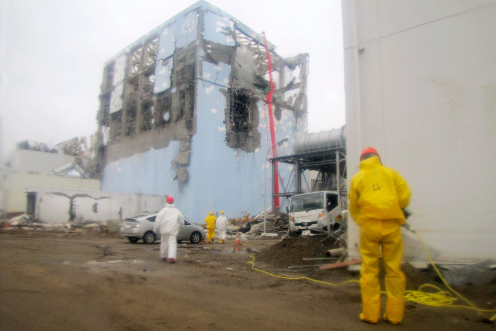 Fotografija prikazuje posljedice nuklearne katastrofe na nuklearnoj elektrani Fukushima Daiichi na japanskoj obali Tihog oceana. Fotografija prikazuje oštećenja na zgradi u kojoj se nalazi nuklearni reaktor. Vide se veliki otvori nastale pod utjecajem plimnoga vala. Ispred zgrada nalaze se radnici odjeveni u zaštitnu opremu, u prvom planu je radnik u žutom zaštitnom odijelu., u drugom planu je radnik u bijelom zaštitnom odijelu.