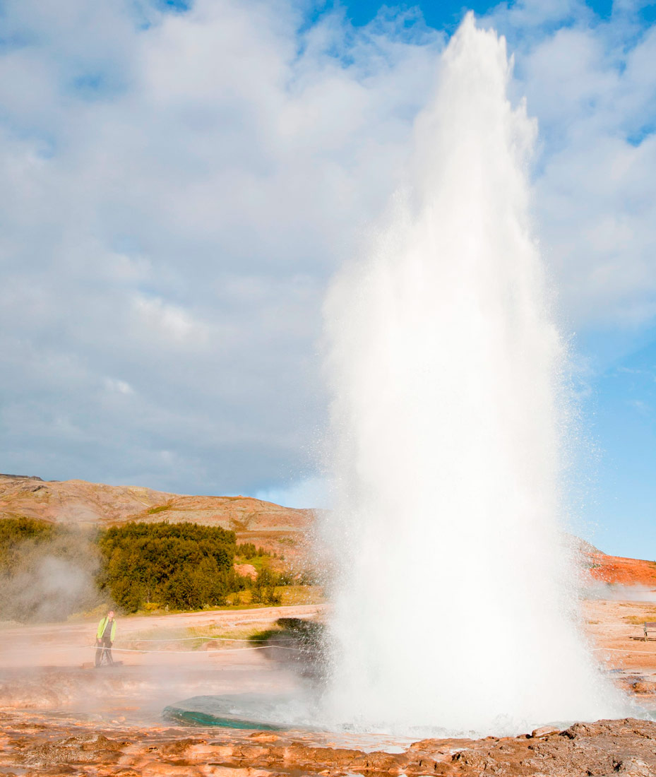 Fotografija prikazuje erupciju gejzira, iz otvora u stjenovitom tlu izdiže se bijeli vodoskok vode i vruće pare.