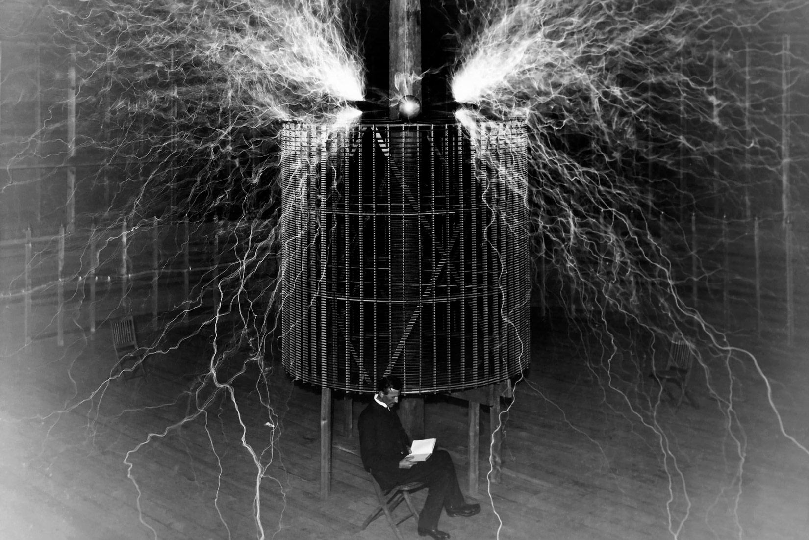 Fotografija prikazuje slavnog fizičara Nikolu Tesla. Fizičar, odjeven u crno odijelo sjedi ispred kaveza s elektromotorom, u rukama drži knjigu. Iznad elektromotora sijevaju bijele iskre različitih duljina, pojedine sežu do tla.