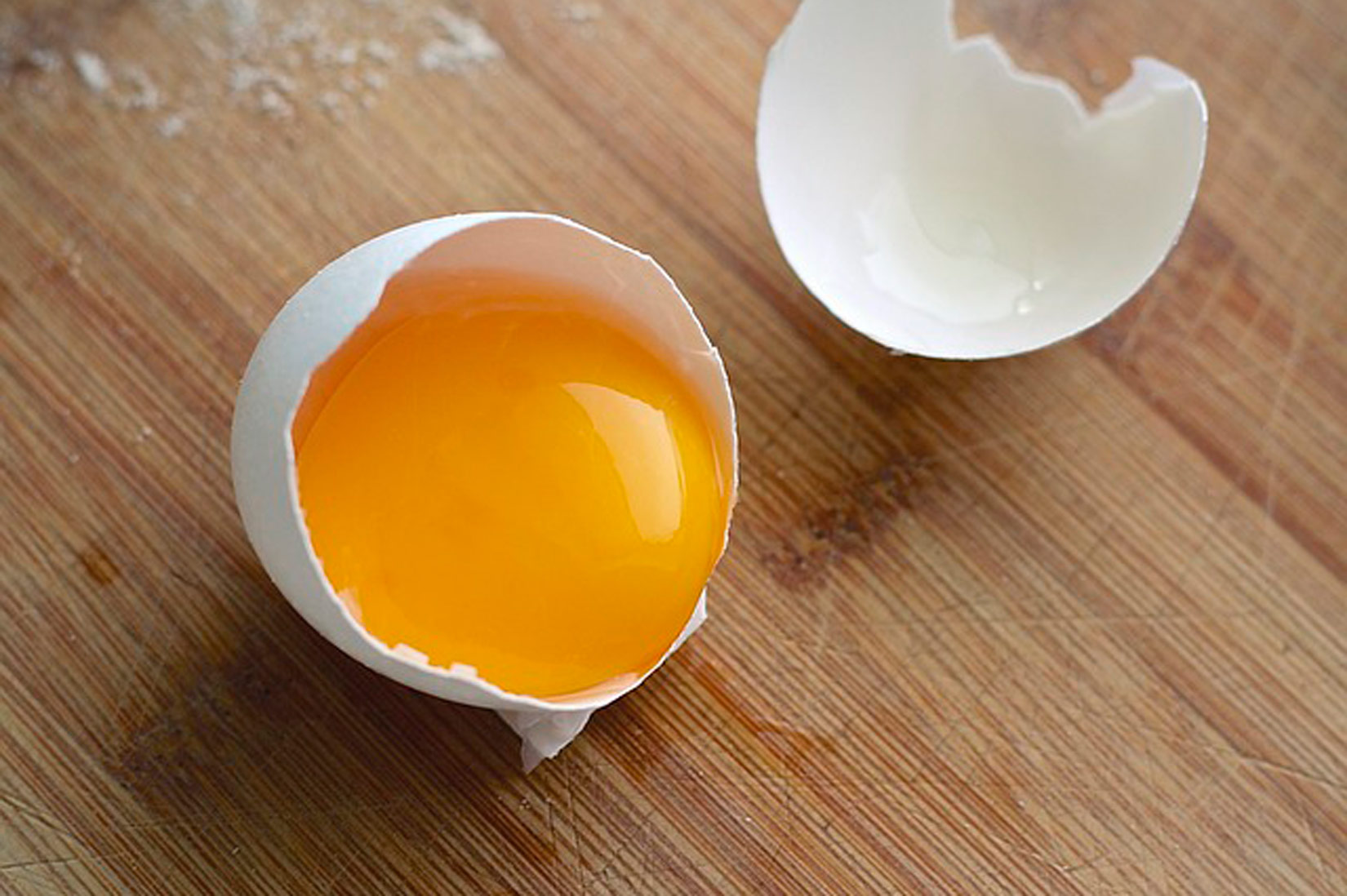 Na slici je razbijeno jaje u krupnom planu. Vide se dva dijela ljuske, jedan je prazan, a u drugom je žumanjak.