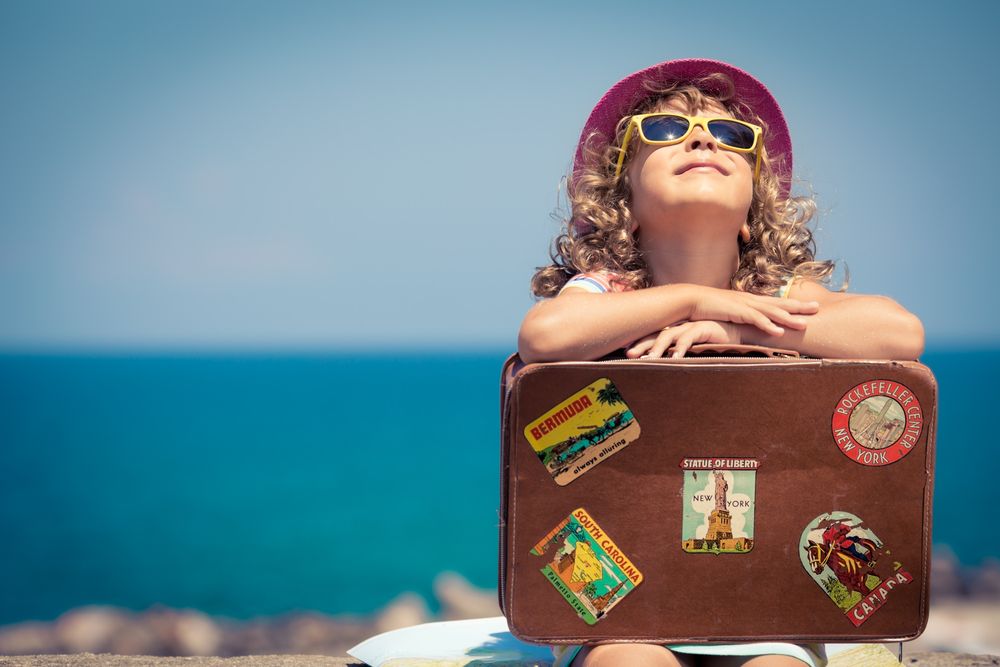 Fotografija prikazuje djevojčicu koja drži kovčeg, a u pozadini se vidi more.