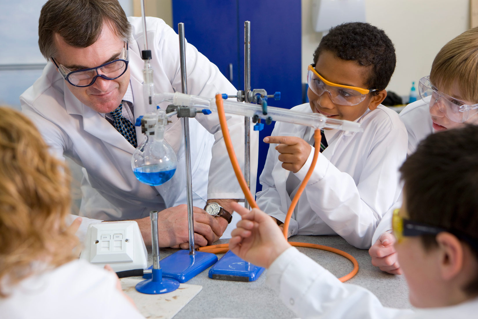 Na slici je prikazana aparatura za destilaciju u kemijskom laboratoriju- učionici. Učenici okupljeni oko svoga učitelja pokazuju prstom što se zbiva tijekom procesa. Na slici su prikazana tri učenika i učenica s leđa.Svi nose zaštitnu opremu (bijele kute, naočale ).