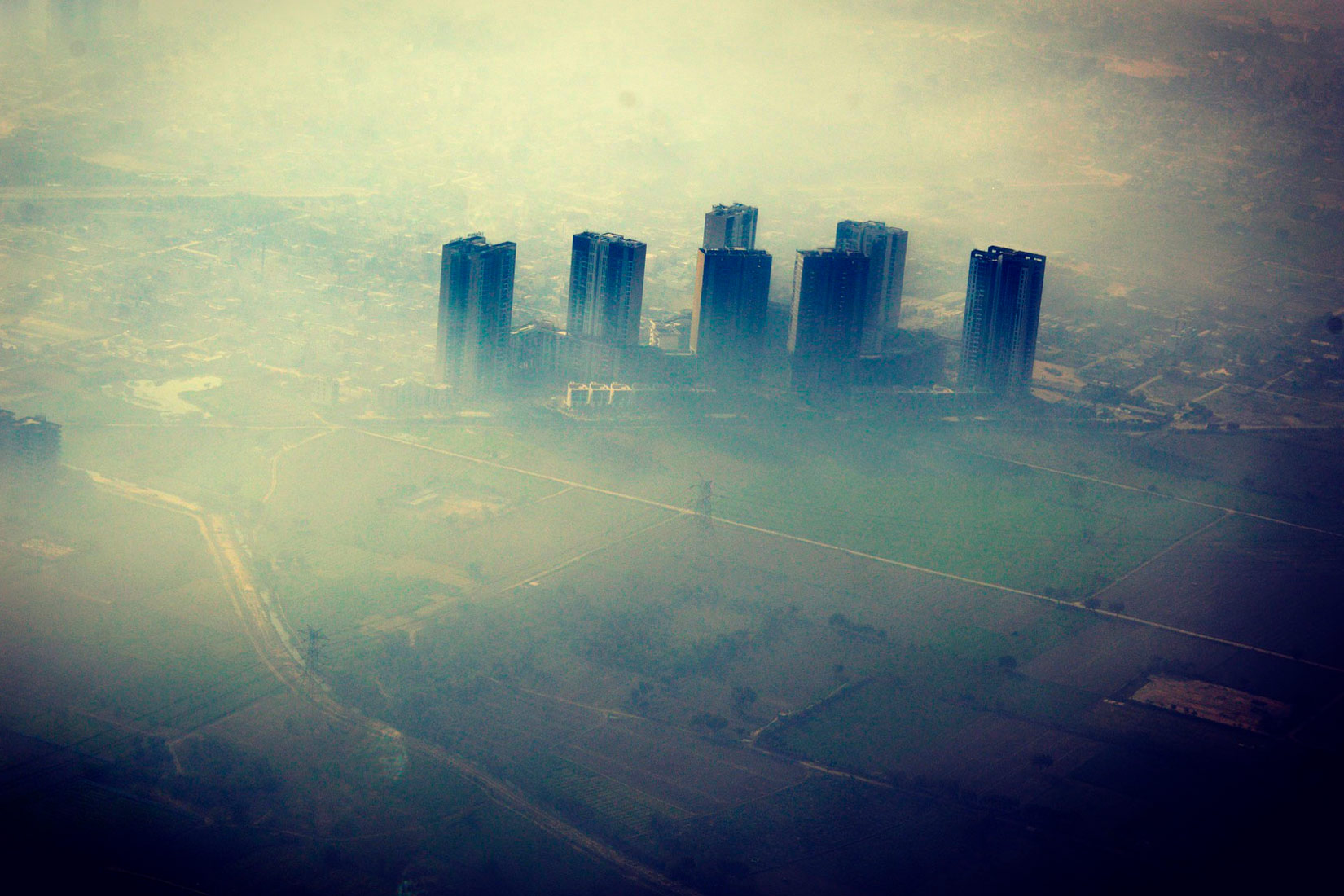 Slika prikazuje grad iz ptičje perspektive. Prekriven je sivo bijelom izmaglicom. U daljini se vidi šest visokih zgrada.