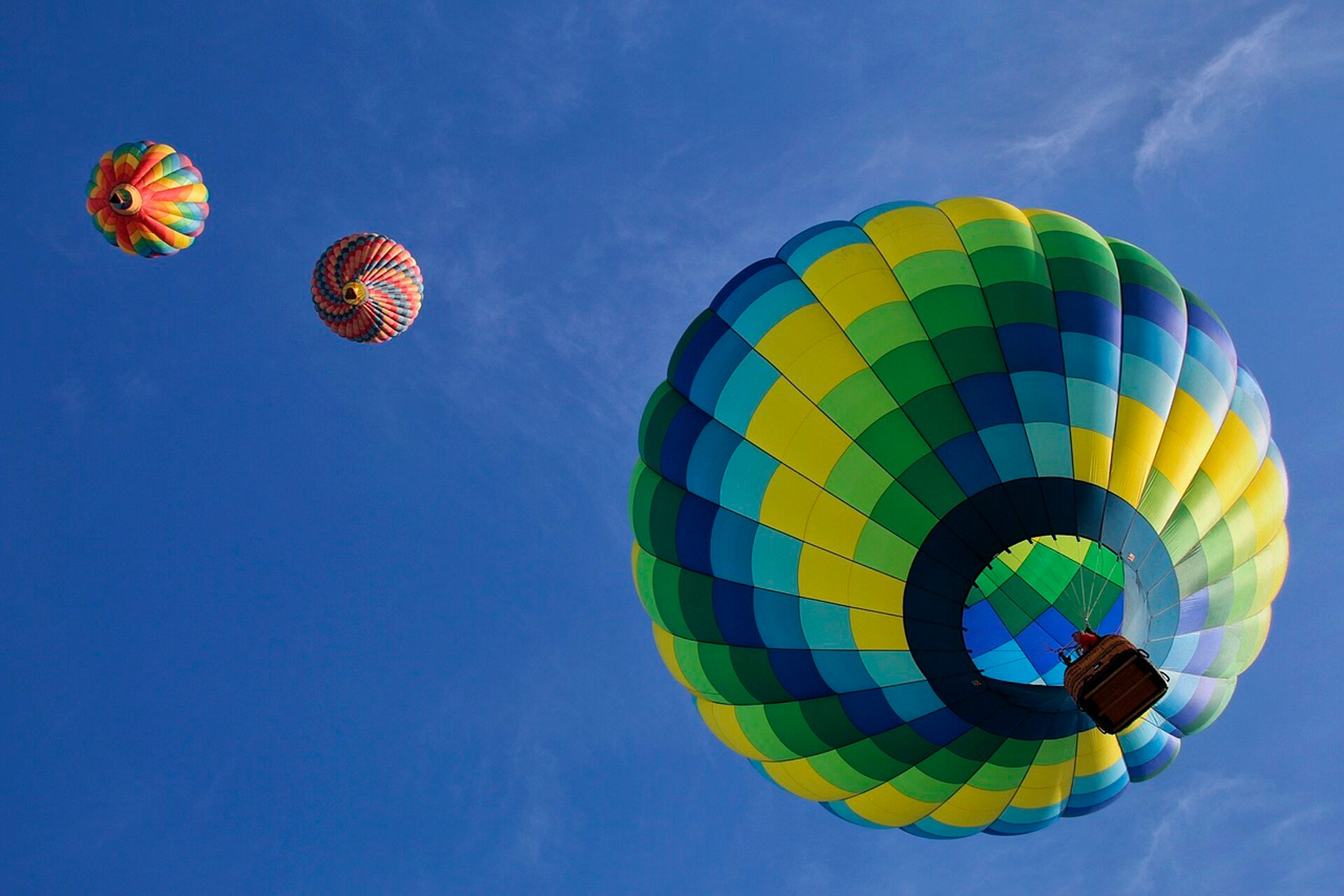 Balon na vrući zrak je prikazan u plavoj, zelenoj i žutoj boji. Ispod balona je pričvršćena košara užadima. Balon se digao u visinu prema plavom nebu.