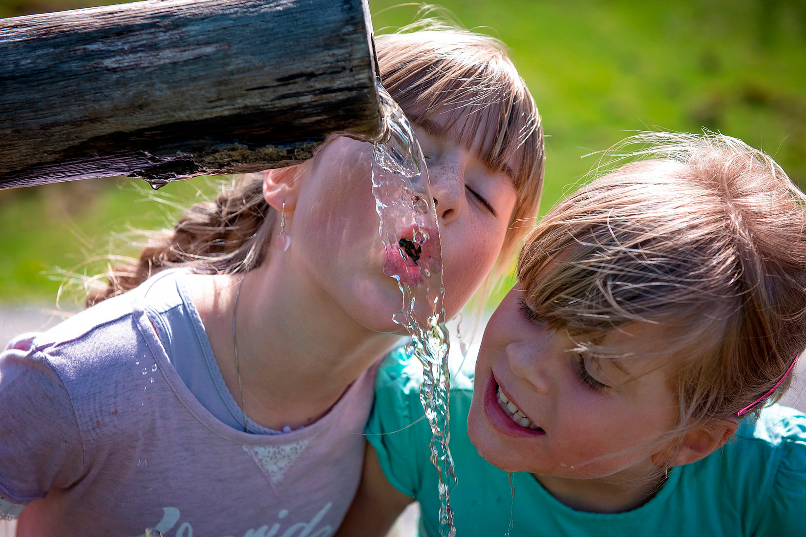 Na fotografiji su prikazane dvije djevojčice kako pokušavaju piti vodu iz cijevi koja se nalazi u visini njihovih glava.Voda je izvor života.