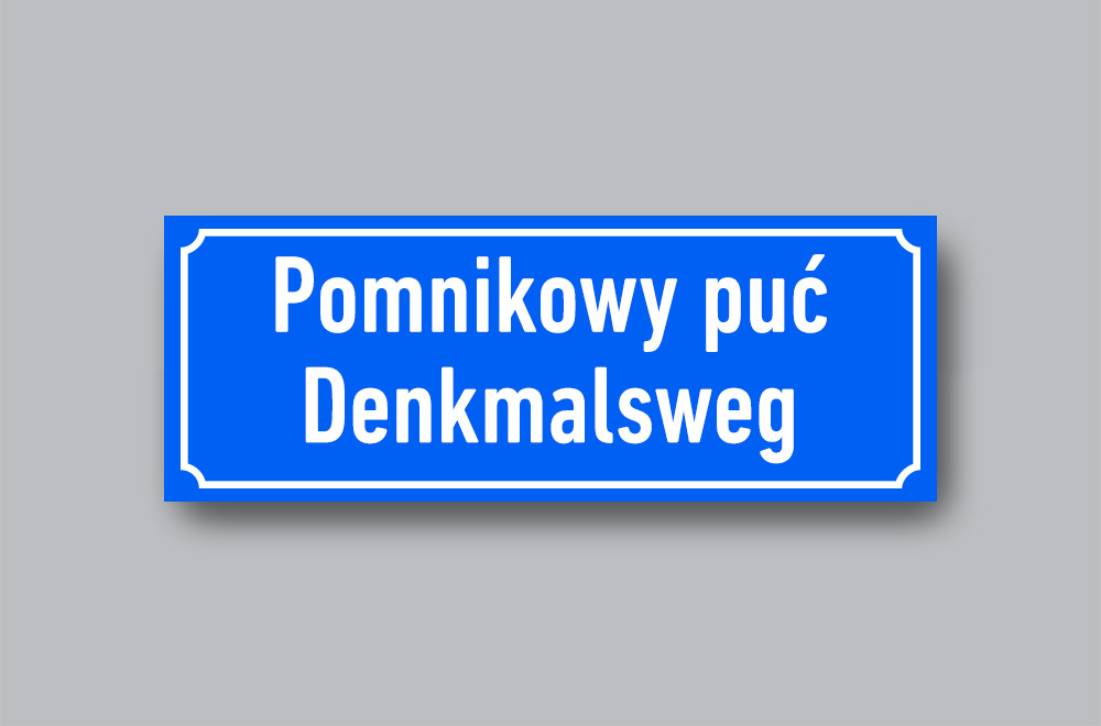Slika prikazuje cestovnu oznaku mjesta na njemačkome i jeziku Lužičkih Srba Pomnikowy puć / Denkmalsweg.