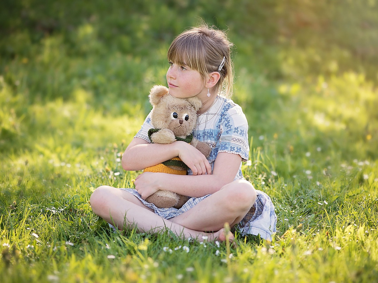 Djevojčica sjedi na livadi i grli igračku medvjedića.