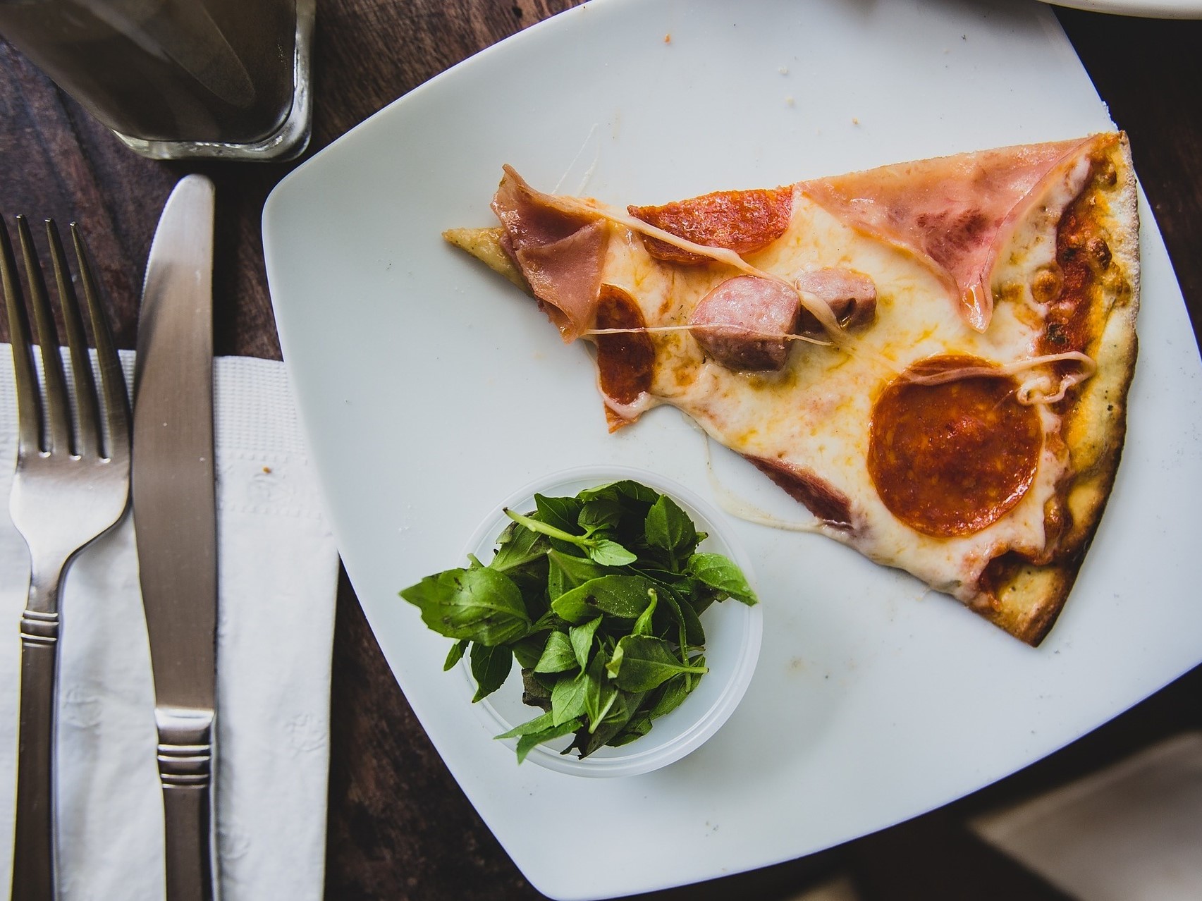 Bijeli četvrtasti tanjur na kojem se nalazi trokut pizze i začinsko bilje u maloj posudi. S lijeve strane tanjura je ubrus s nožem i vilicom te čaša.