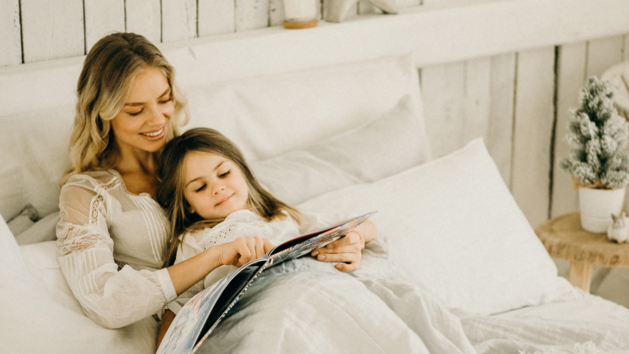 Majka i djevojčica leže u krevetu i zajedno čitaju slikovnicu. 