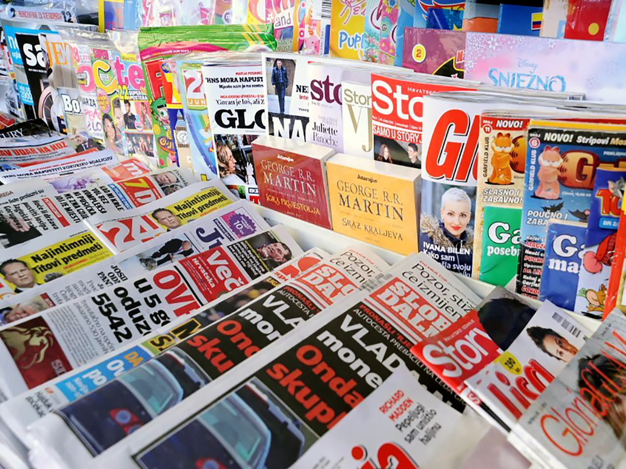 Brojne dnevne i tjedne tiskovine poslagane na polici kioska. 
