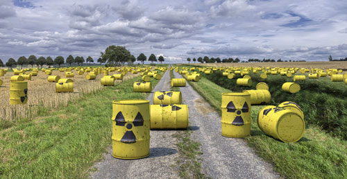 Fotokgrafija prikazuje nuklearni otpad smješten u žute bačve s crniom oznakom radioaktivnosti.Bačve su raspoređene po livadi koja ima neasfaltirani puteljak.Nebo je oblačno.