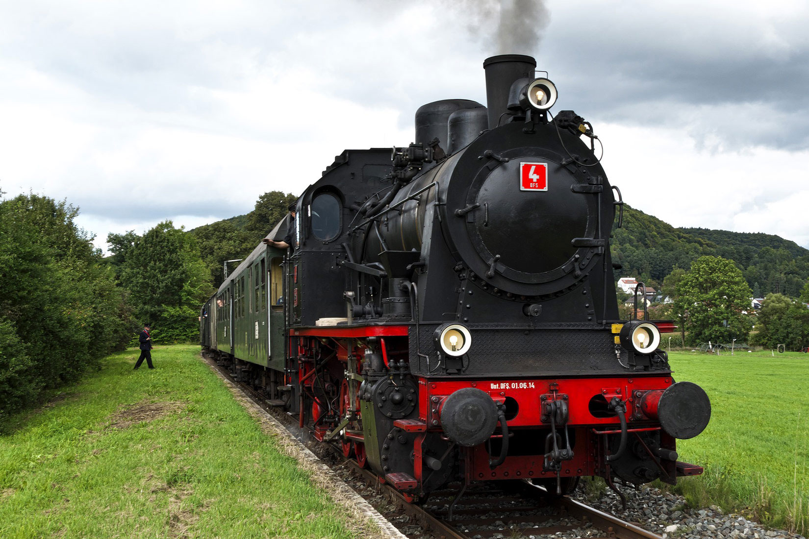 Fotografija prikazuje parnu lokomotivu , uslikanu s prednje strane. Lokomotiva je crno-crvene boje. Putuje kroz krajobraz u kojem se vidi livada i stabla.